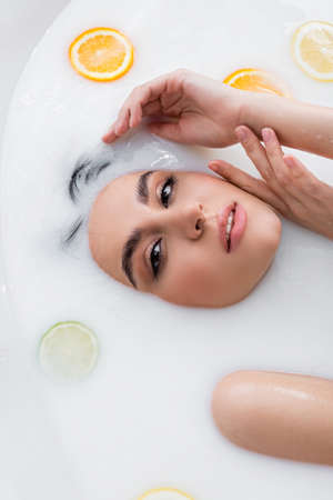 pretty woman holding hands near face and looking at camera while relaxing in milk bath with citrusesの写真素材