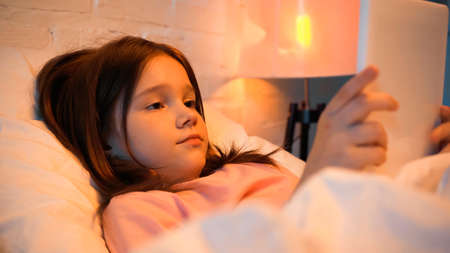 Preadolescent girl using digital tablet near blurred blanket on bed