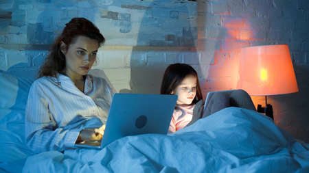 Freelancer using laptop near daughter with digital tablet on bed