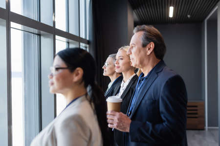 Side view of businesswoman looking at window near colleagues on blurred foregroundの写真素材