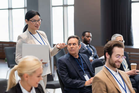 Smiling asian businesswoman with laptop talking near business people during seminarの写真素材