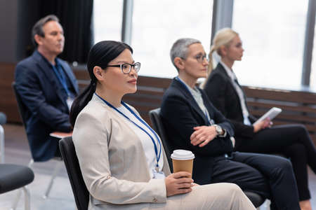 Asian businesswoman with coffee to go sitting near colleagues in conference roomの写真素材