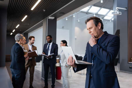 Mature businessman looking at laptop near interracial colleagues on blurred background