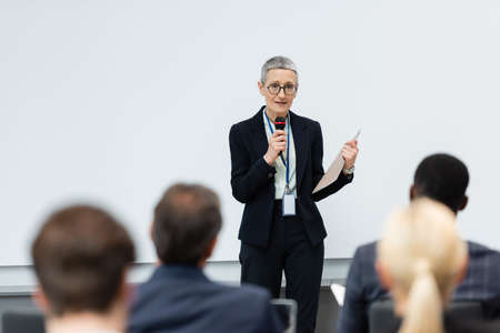 Mature speaker with paper folder talking near blurred business people during meeting