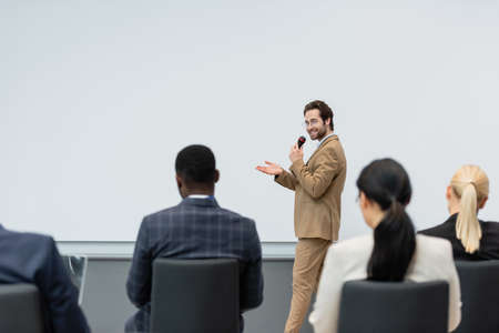 Smiling speaker talking and pointing with hand near blurred multiethnic business people during seminarの写真素材