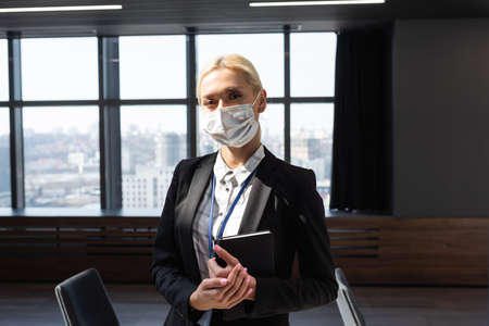 blonde businesswoman in medical mask standing with notebook in officeの写真素材