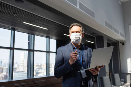 mature businessman in medical mask looking away while holding laptop in office