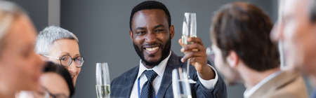 smiling african american businessman holding champagne glass near colleague on blurred foreground, bannerの写真素材