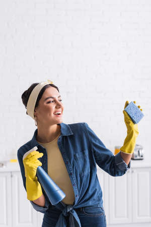 Cheerful housewife with sponge and spray bottle in kitchen