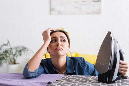 Displeased woman holding blurred iron near clothes on boardの写真素材