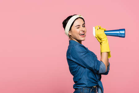 Smiling housewife in rubber gloves holding detergent and winking at camera isolated on pinkの写真素材