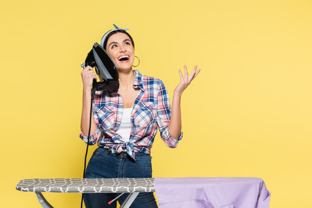 Cheerful woman holding iron near ear and board isolated on yellowの写真素材