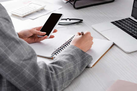 Cropped view of businesswoman with smartphone writing on notebook near laptop and blurred calculator