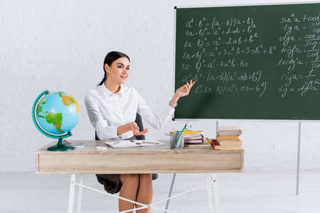 Smiling teacher pointing at chalkboard near globe and books in classroomの写真素材
