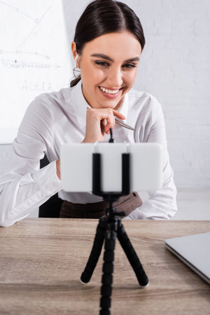 Cheerful businesswoman in earphone having video chat on blurred smartphone in officeの写真素材