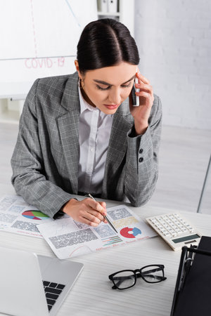 Businesswoman looking at charts on document and talking on smartphone in officeの写真素材