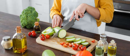 cropped view of woman in apron cutting zucchini near cherry tomatoes on chopping board, banner