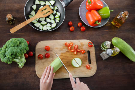 top view of woman cutting zucchini near sliced cherry tomatoes on chopping board and frying pan on tableの写真素材