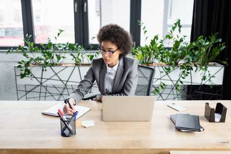 African american businesswoman in eyeglasses writing on notebook near laptop and smartphoneの写真素材
