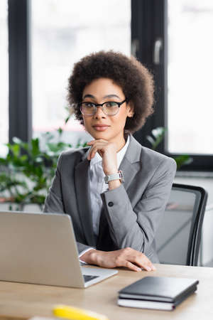 African american businesswoman sitting near blurred laptop and notebooksの写真素材