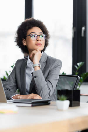 Dreamy african american manager looking away near blurred working tableの写真素材