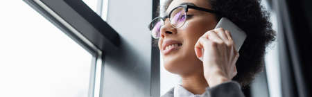 Low angle view of african american manager in eyeglasses talking on mobile phone near office window,...