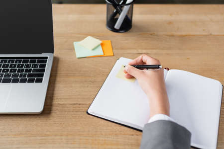 Cropped view of businesswoman holding pen near notebook with sticky note and laptopの写真素材