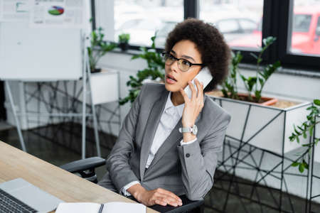 African american manager talking on smartphone near blurred laptop and notebookの写真素材