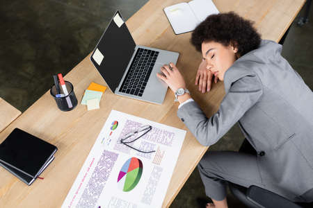 High angle view of african american businesswoman lying with closed eyes near laptop and paper on...