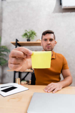 young man showing empty sticky note while working at home on blurred backgroundの写真素材