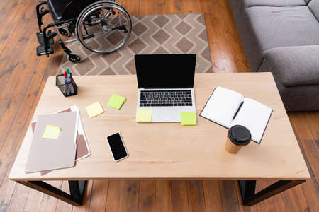 high angle view of desk with gadgets, documents and takeaway drink near wheelchair