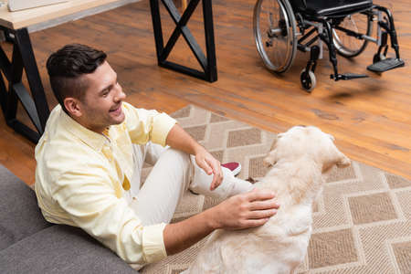 smiling, handicapped man stroking labrador while sitting on floor near wheelchairの写真素材