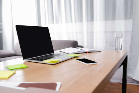 laptop and smartphone with blank screen near glass of water, sticky notes and notebook on desk