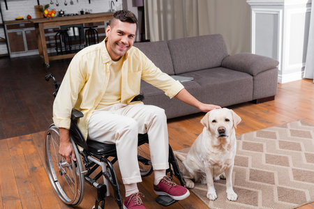 handicapped man petting labrador dog while sitting in wheelchair at homeの写真素材