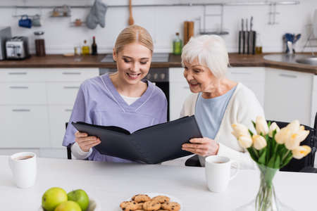 pleased senior woman and nurse looking at photo album in kitchenの写真素材