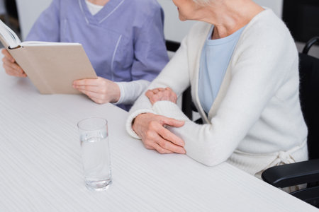 partial view of nurse reading book to aged woman near glass of water, blurred backgroundの写真素材