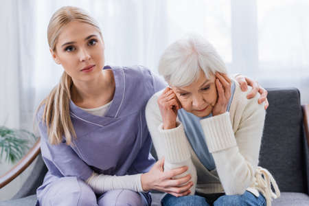 young nurse looking at camera while embracing senior woman sitting with bowed head