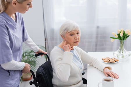 disabled senior woman pointing at hearing aid while sitting in wheelchair near social workerの写真素材