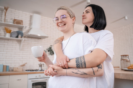 brunette woman hugging tattooed girlfriend with cup of coffee in kitchenの写真素材