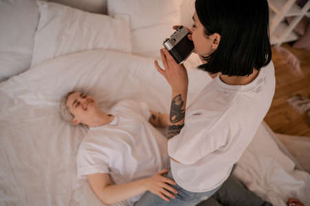tattooed woman taking photo of blurred girlfriend lying on bed and smiling on retro camera