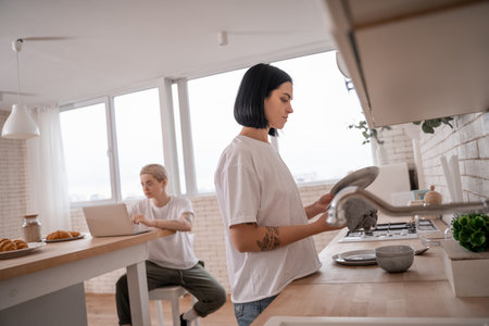 brunette woman washing plate near girlfriend using laptop on blurred backgroundの写真素材