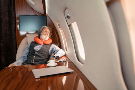 Mature businessman in neck pillow sleeping near laptop and coffee in plane