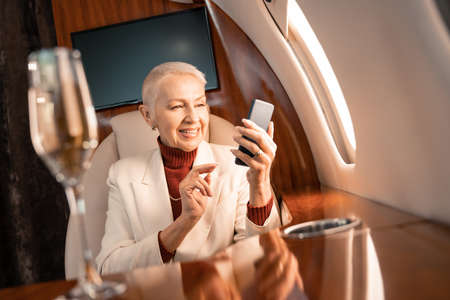 Smiling businesswoman using smartphone near blurred glass of champagne in plane