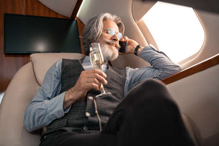 Low angle view of smiling businessman talking on smartphone and holding champagne in planeの写真素材