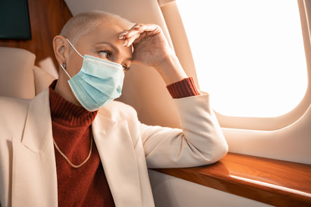 Mature businesswoman in medical mask looking at window in private planeの写真素材