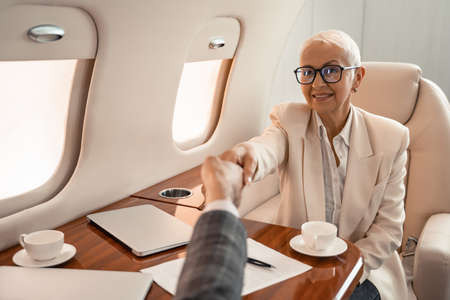 Smiling businesswoman shaking hands with businessman near paper and laptops in private jet
