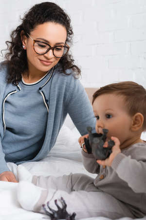 happy african american woman looking at blurred infant boy playing with toy on bed
