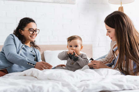 happy interracial women looking at infant boy playing with toy while sitting on bed