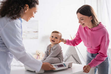 smiling african american pediatrician and mother looking at infant boy on baby scaleの写真素材