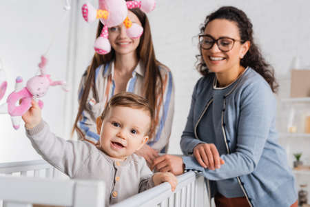 blurred interracial women near happy infant boy in baby cribの写真素材